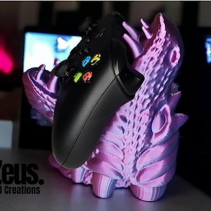 Könnte beinhalten: Ein schwarzer Xbox-Controller liegt auf einem 3D-gedruckten Ständer. Der Ständer ist ein detailliertes, mehrschichtiges Design in Rosa- und Lilatönen. Der Controller hat farbige Knöpfe. Der Text "Zeus. 3D Creations" ist sichtbar.