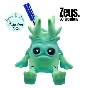 May include: A 3D-printed teal and green monster-like desk organiser with a blue pen. The creature has large black eyes, spiky protrusions, and articulated arms and legs. The text "Zeus. 3D Creations" is visible.