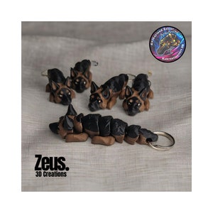 May include: Five German Shepherd dog keychains made from brown and black polymer clay. The keychains are in various poses, including sitting, lying down, and standing. The keychains are attached to silver key rings. The keychains are on a light brown fabric background. The image also includes a small round sticker with the text "Authorized Seller Kekreationsart.com".