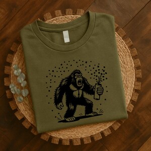 May include: Olive green t-shirt featuring a black graphic of a roaring Bigfoot holding a firecracker. The design includes a burst of confetti-like shapes. The shirt is displayed on a woven, circular placemat.