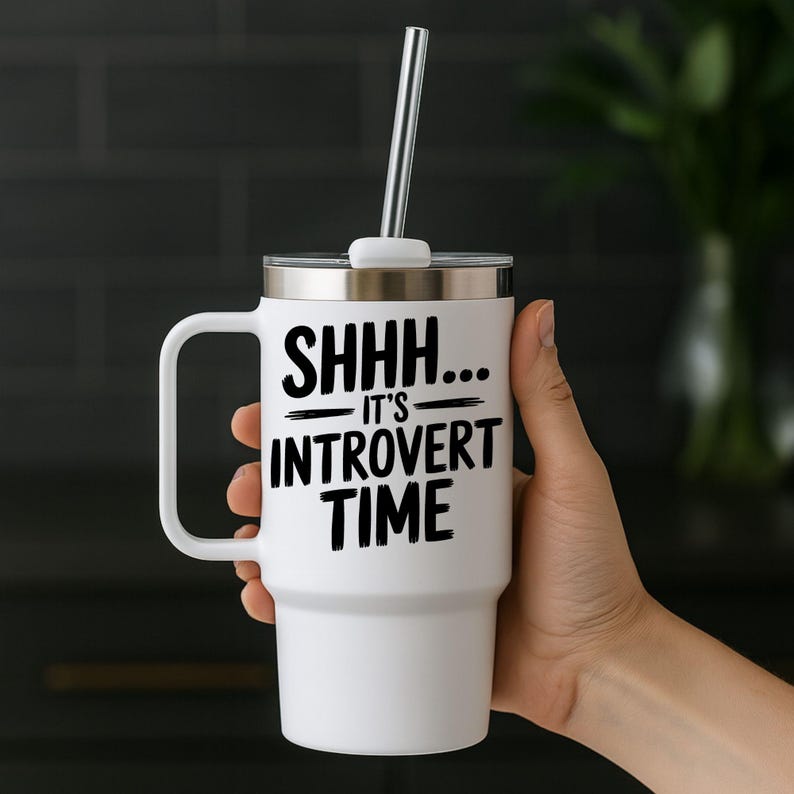 May include: White travel mug with a handle, silver lid, and a metal straw. The mug has the text "SHHH... IT'S INTROVERT TIME" in black. The mug is held by a person.