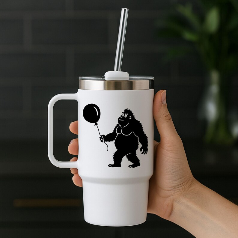 May include: White travel mug with a black graphic of a Bigfoot holding a balloon. The mug has a silver lid, a white handle, and a silver straw. The mug is held by a person's hand.