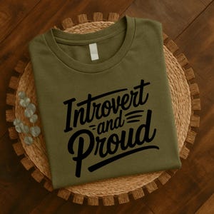 May include: An olive green t-shirt with the words "Introvert and Proud" in a black, cursive font. The shirt is folded and resting on a woven, circular placemat. The background is a wooden surface.
