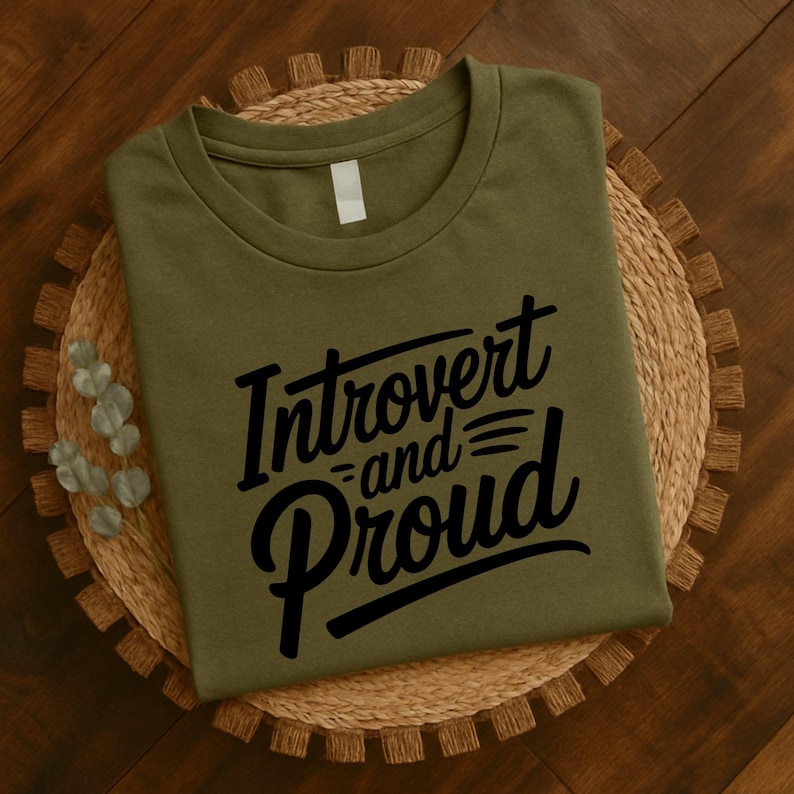 May include: An olive green t-shirt with the words "Introvert and Proud" in a black, cursive font. The shirt is folded and resting on a woven, circular placemat. The background is a wooden surface.
