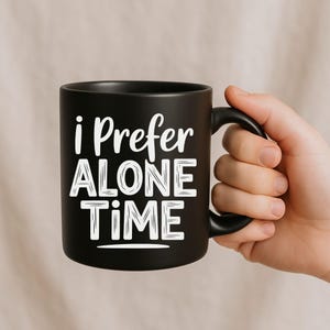 May include: A black ceramic mug with the text "i Prefer ALONE TIME" in white. The mug has a black handle and is held by a hand. The mug is a standard size for coffee or tea.