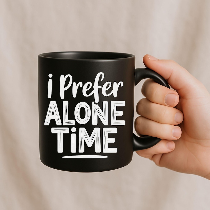 May include: A black ceramic mug with the text "i Prefer ALONE TIME" in white. The mug has a black handle and is held by a hand. The mug is a standard size for coffee or tea.