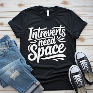 May include: Black t-shirt with the white text "Introverts need Space." The shirt is paired with blue jeans and black canvas sneakers with white laces and soles. The items are arranged on a light wood-grain surface.