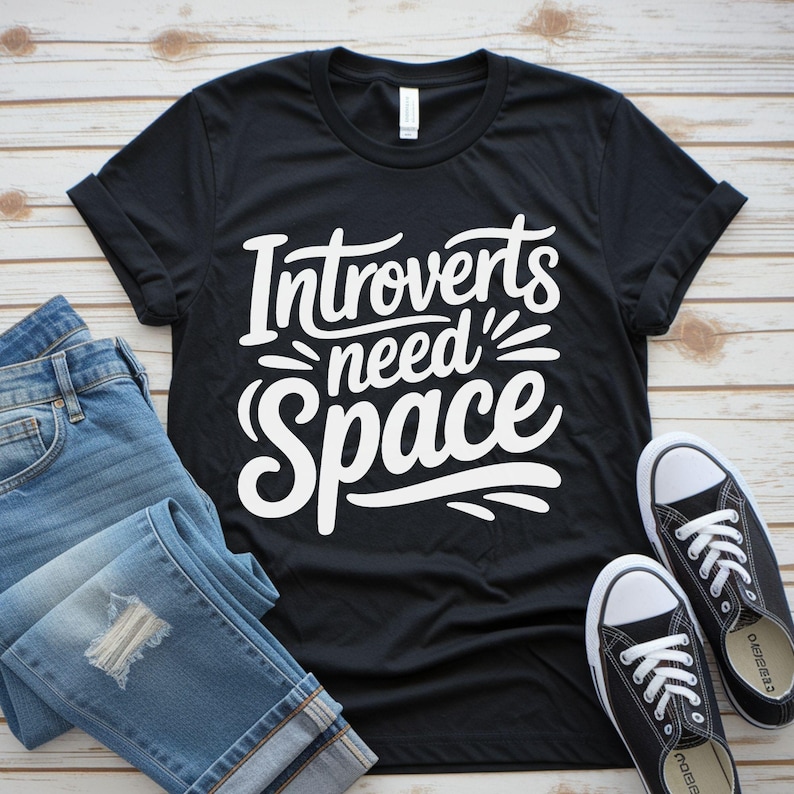 May include: Black t-shirt with the white text "Introverts need Space." The shirt is paired with blue jeans and black canvas sneakers with white laces and soles. The items are arranged on a light wood-grain surface.