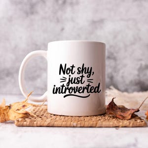 May include: White ceramic mug with the text "Not shy, just introverted" in black script. The mug has a curved handle and sits on a woven mat with dried leaves.