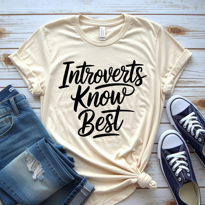 May include: Cream-colored t-shirt with the text "Introverts Know Best" in black script. The shirt is paired with blue jeans and navy blue canvas sneakers. The shirt is knotted at the waist.