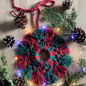 以下が含まれることがあります： フリンジの縁取りが施された赤と緑の糸で編まれたリース。リースは松の枝と松ぼっくりで飾られています。