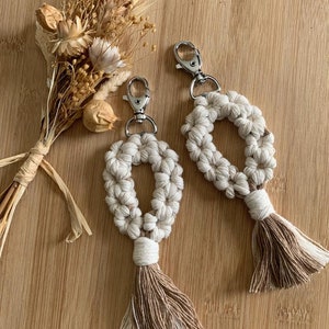 May include: Two beige macrame keychains with brown tassels and silver key rings. The keychains are in the shape of an oval with a hole in the center.