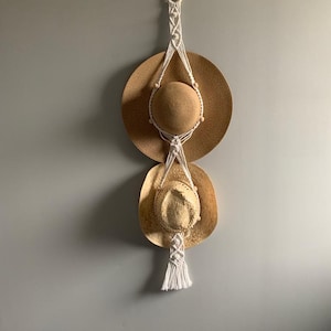 May include: White macrame hat holder with three straw hats hanging from it. The holder is attached to a wall with a wooden ring.