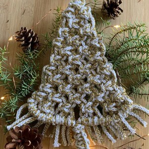Può includere: Un ornamento per albero di Natale in macramé bianco e oro con frange. L'ornamento è circondato da rami di pino e pigne.