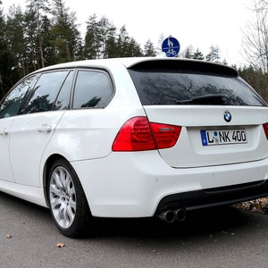 May include: A white BMW 3 Series wagon with a license plate that reads "L-NK 400". The car is parked on a paved road with trees in the background.