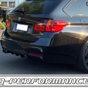 May include: Black BMW wagon with a custom rear bumper and exhaust system. The car has black wheels and a black paint job. The rear bumper has a diffuser and four exhaust tips. The car is parked on a paved surface.