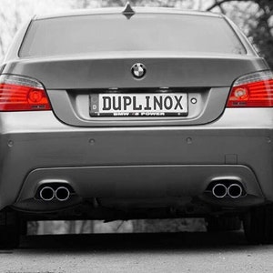 May include: Rear view of a silver BMW with a black number plate that reads "DUPLINOX" with the text "BMW IS POWER" below. The car has dual exhaust pipes and red tail lights. The image is in black and white except for the red tail lights and the number plate text.
