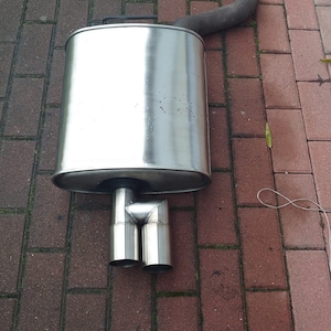 Peut inclure: Un silencieux d'échappement de voiture en acier inoxydable avec deux embouts d'échappement ronds. Le silencieux est posé sur une surface en briques rouges.