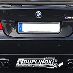 May include: Rear view of a black BMW M5 with red taillights. The car has a license plate, the BMW logo, and the M5 emblem. A Duplinox performance exhaust systems sticker is visible.