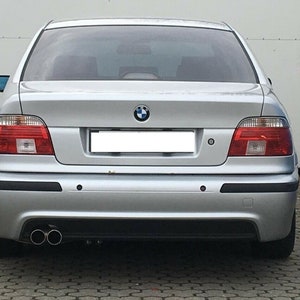 Könnte beinhalten: Ein silberner BMW 5er Limousine mit schwarzem Stoßfänger und zwei Auspuffrohren. Das Auto hat ein weißes Nummernschild und ein BMW-Logo auf dem Kofferraum.