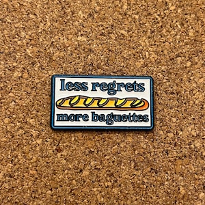Peut inclure: Une broche émaillée rectangulaire avec une bordure bleu foncé et un fond blanc. Le texte less regrets est écrit en lettres bleues au-dessus d'un dessin de baguette dorée, avec le texte more baguettes en lettres bleues sous le dessin.