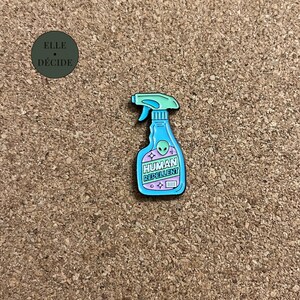 Peut inclure: Épingle en émail fantaisie en forme de flacon pulvérisateur, avec une palette de couleurs bleu sarcelle et bleu clair. L'épingle présente un motif d'extraterrestre et les mots "HUMAN REPELLENT". L'épingle est posée sur un fond en liège brun.