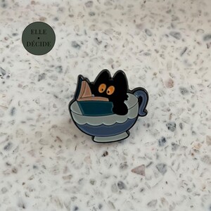 Peut inclure: Une broche en émail métallique représentant un chat noir aux grands yeux orange assis dans une tasse à thé bleue, lisant un livre ouvert à couverture sarcelle. La broche a un contour sombre et une finition brillante. Le texte ELLE DÉCIDE est visible.
