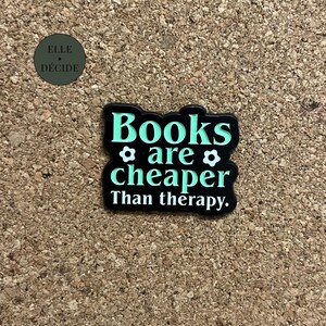 Peut inclure: Épinglette noire et turquoise avec le texte "Books are cheaper than therapy". L'épinglette a une bordure noire et des lettres turquoise. L'épinglette est sur une surface en liège marron. L'épinglette a également deux petites fleurs.