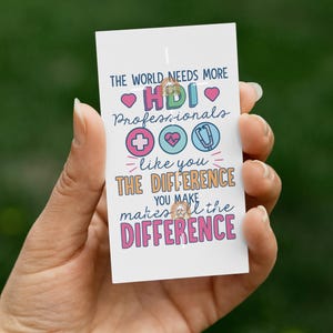May include: A white card with colorful text and graphics is held in a hand. The text reads "THE WORLD NEEDS MORE HDI Professionals" with a heart, medical cross, heart, and stethoscope icons. The card also says "like you THE DIFFERENCE YOU MAKE makes all the DIFFERENCE."