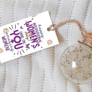 May include: White gift tag with purple text that reads "Happy International Women's Day! Shine on, you amazing women!" with a gold glitter background and a butterfly graphic. The tag is tied to a clear glass globe with a cork top and twine.