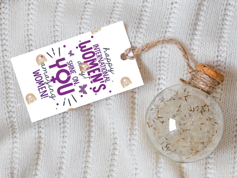 May include: White gift tag with purple text that reads "Happy International Women's Day! Shine on, you amazing women!" with a gold glitter background and a butterfly graphic. The tag is tied to a clear glass globe with a cork top and twine.