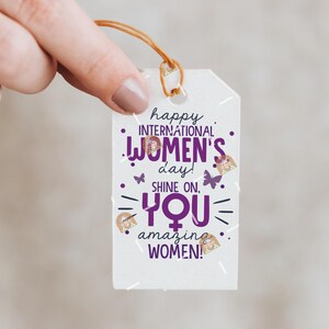 May include: White gift tag with purple text that reads "happy international women's day! shine on, you amazing women!" with gold glitter accents and butterfly graphics.