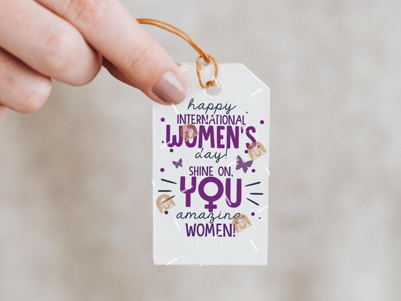 May include: White gift tag with purple text that reads "happy international women's day! shine on, you amazing women!" with gold glitter accents and butterfly graphics.
