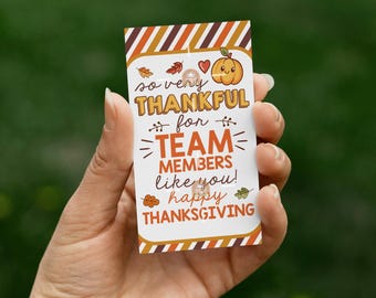 May include: A Thanksgiving-themed gift tag held in a hand. The tag features a pumpkin illustration, fall leaves, and the text "so very THANKFUL for TEAM MEMBERS like you! happy THANKSGIVING" in a mix of fonts and colors.
