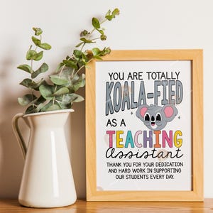 May include: A framed print with the text "YOU ARE TOTALLY KOALA-FIED AS A TEACHING ASSISTANT." A cartoon koala is featured. A white vase with green sprigs is to the left of the frame. The frame is light brown.