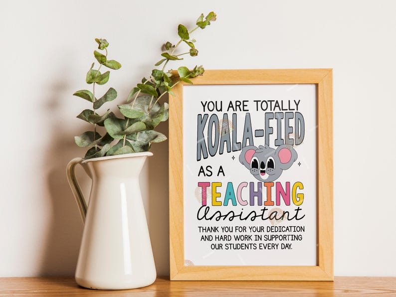 May include: A framed print with the text "YOU ARE TOTALLY KOALA-FIED AS A TEACHING ASSISTANT." A cartoon koala is featured. A white vase with green sprigs is to the left of the frame. The frame is light brown.