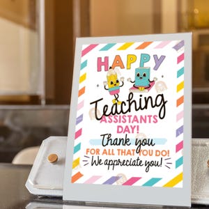May include: A colorful sign with the text "HAPPY TEACHING ASSISTANTS DAY!" and a thank you message. The sign features cartoon illustrations of a pencil and a book. The sign is displayed in a silver stand.