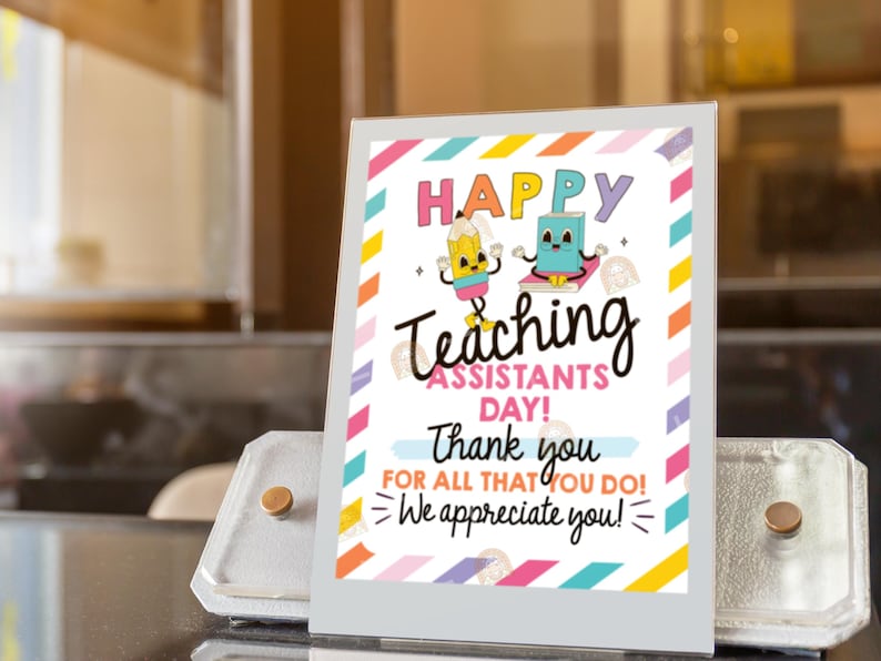 May include: A colorful sign with the text "HAPPY TEACHING ASSISTANTS DAY!" and a thank you message. The sign features cartoon illustrations of a pencil and a book. The sign is displayed in a silver stand.