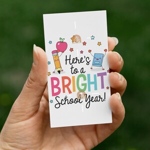 May include: A rectangular white card featuring the text Here's to a BRIGHT School Year! in black and colorful letters. The design includes cartoon illustrations of a pencil, an apple, a book, and small stars in various colors.