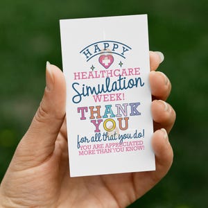 May include: A white card with colorful text is held in a hand against a blurred green background. The card reads "HAPPY HEALTHCARE Simulation WEEK! THANK YOU for all that you do! YOU ARE APPRECIATED MORE THAN YOU KNOW!"