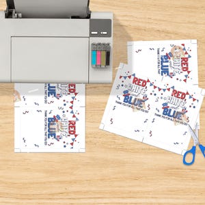 May include: A printer printing patriotic-themed cards with the words "Red, White, and Blue" and "Thank You For All That You Do." The cards have a festive design with stars and bunting. Blue scissors are on the wooden surface.