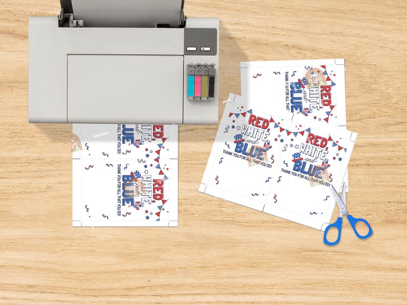 May include: A printer printing patriotic-themed cards with the words "Red, White, and Blue" and "Thank You For All That You Do." The cards have a festive design with stars and bunting. Blue scissors are on the wooden surface.