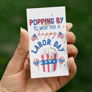Puede incluir: Una tarjeta blanca con el texto "Popping by to wish you a happy Labor Day" en rojo y azul. La tarjeta presenta un cubo de palomitas de maíz con los colores de la bandera estadounidense y dos globos. También hay dos pequeñas banderas americanas.