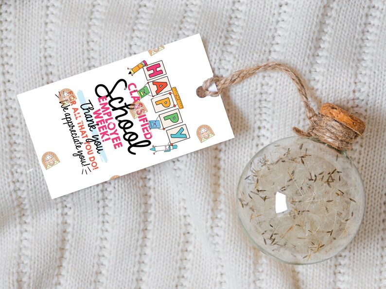 May include: A clear glass ornament filled with dried plant material, attached to a tag with the words "HAPPY SCHOOL EMPLOYEE WEEK!" The tag is white with colorful illustrations and a brown twine string.