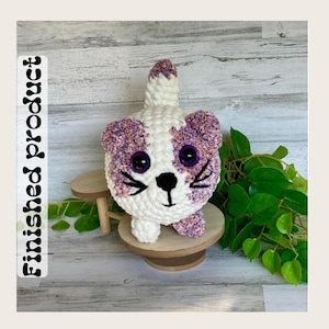 May include: A handmade crochet cat toy in white and purple. The cat has large purple eyes, black whiskers, and a black nose. The text "Finished product" is printed vertically on the left side of the image. The cat is sitting on a small wooden stand.