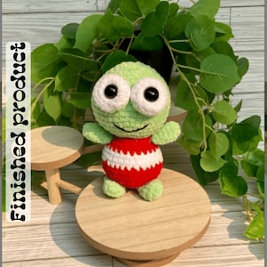 May include: Green crocheted frog amigurumi toy wearing a red and white striped outfit. The toy is sitting on a wooden stand.