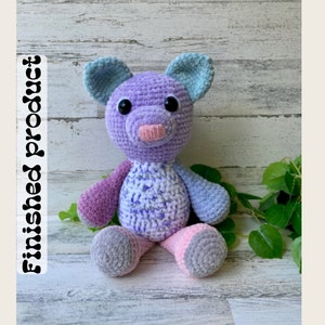 May include: A crocheted plush toy in the shape of a pig. The pig is purple with pink ears, a pink nose, and black eyes. The pig is sitting on a white wooden surface.
