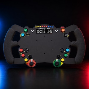 May include: A black Fanatec racing steering wheel with textured grips, featuring a central digital display showing race data, a row of colored LED lights, and multiple multi-colored push buttons and rotary dials arranged on the front face.