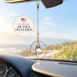 Puede incluir: Un adorno de coche de metal dorado que cuelga de una cadena delicada, con dos anillos circulares entrelazados y dos colgantes de cristal transparente facetado. El cristal superior es esférico y el cristal inferior tiene forma de lágrima.