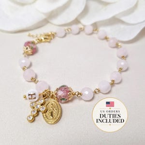May include: A gold-toned bracelet featuring pale pink faceted beads, two floral patterned beads with gold caps, a white bead with the letter H, a gold cross charm with clear stones, and a gold oval medal depicting a religious figure.